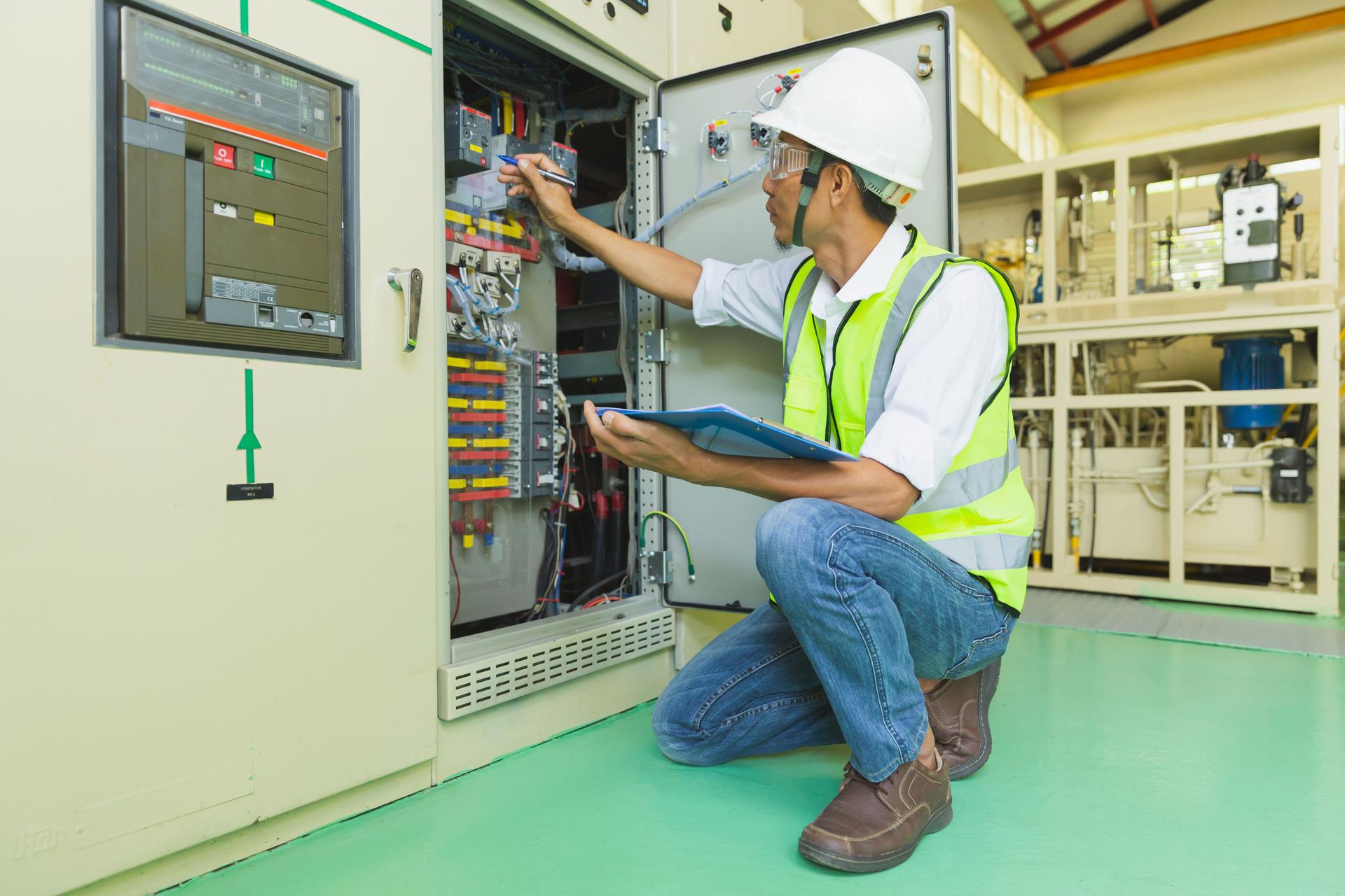 A helmeted technician holds a clipboard while checking and taking notes on the electrical control cabinet.